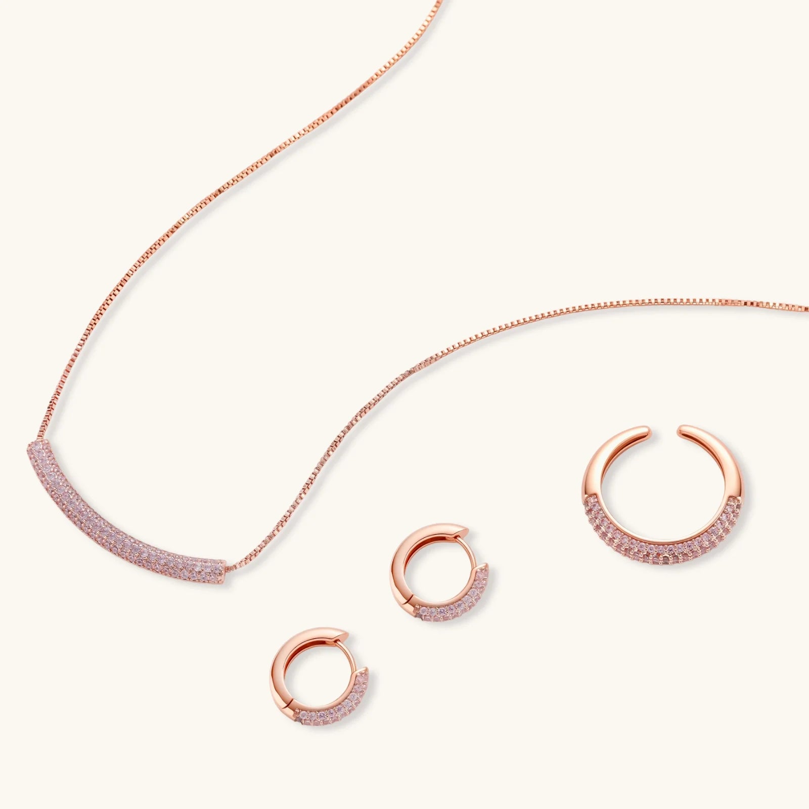 Rose gold pavé zirconia bar necklace, hoop earrings, and open ring set with pink stones on cream background.