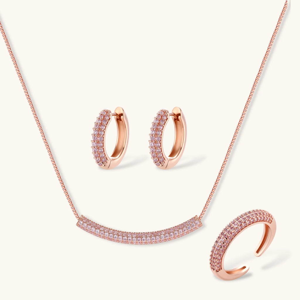 Rose gold pavé zirconia bar necklace, hoop earrings, and open ring set with pink stones on cream background.