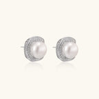 Rhodium-plated sterling silver stud earrings with round freshwater pearls surrounded by geometric crystal halos on cream background.